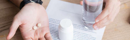 Cropped view of man holding glass of water and pills near business contract on wooden background,...