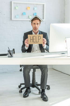 Full length of disappointed bankrupt with notebook on head, holding cardboard with help lettering,...