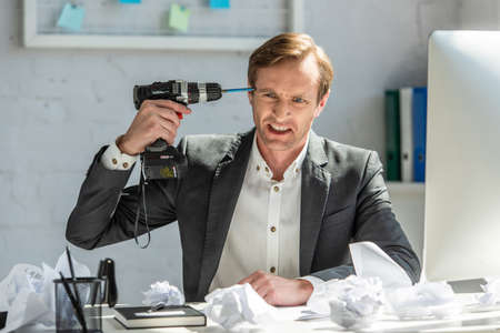 Aggressive office worker holding drill near head, while sitting at workplace with crumpled papers on...