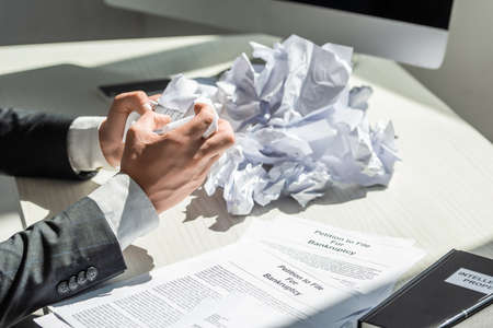 Cropped view of businessman holding crumbled paper near petitions for bankruptcy at workplace on blurred backgroundの写真素材