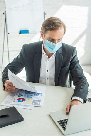 Businessman in medical mask, holding papers and typing on laptop, while sitting at workplace on blurred backgroundの写真素材