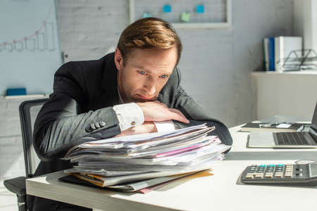 Disappointed businessman leaning on pile of papers, while sitting at workplace on blurred backgroundの写真素材