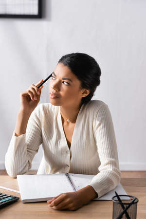 Thoughtful african american woman holding pen and looking away while sitting at desk in office on blurred backgroundの写真素材