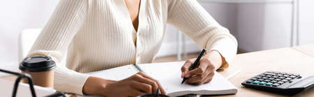 Cropped view of african american woman writing on blank notebook while sitting at desk on blurred background, bannerの写真素材
