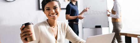 Cheerful african american woman with paper cup looking at camera with blurred office workers on background, bannerの写真素材