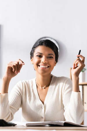 Happy african american businesswoman with pen looking at camera while sitting at desk in office on blurred backgroundの写真素材