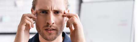 Exhausted young adult man looking at camera and touching head with hands on blurred background,...