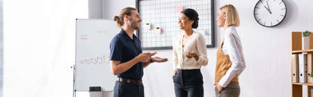 Smiling multicultural office workers talking while standing near mesh organizer with blank stickers and flipchart, bannerの写真素材