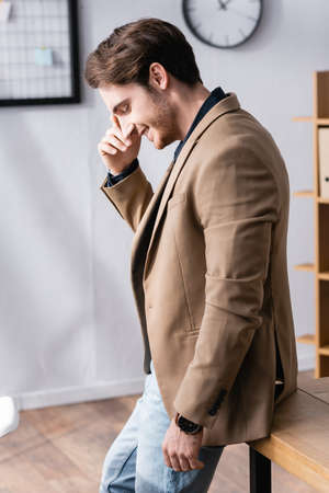 Side view of thoughtful man with closed eyes smiling while leaning on desk in office with on blurred backgroundの写真素材