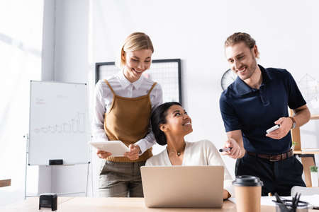 Happy office workers standing near african american woman sitting at table with laptop at workplace