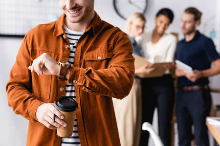 smiling businessman holding coffee to go and looking at wristwatch near colleagues talking on...