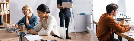 Man holding laptop and standing near multicultural colleagues sitting at workplaces in office,...