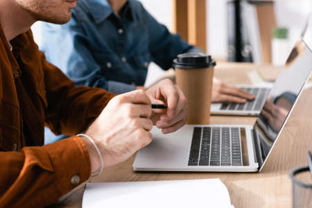 Cropped view of man with pen looking at blank screen of laptop, while sitting at workplace with blurred woman on backgroundの写真素材