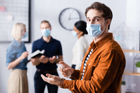 young manager in medical mask holding disinfectant while looking at camera near businesspeople on...