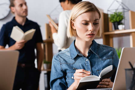 Skeptical blonde woman writing on notebook while looking at laptop in office with blurred multicultural co-workers on backgroundの写真素材