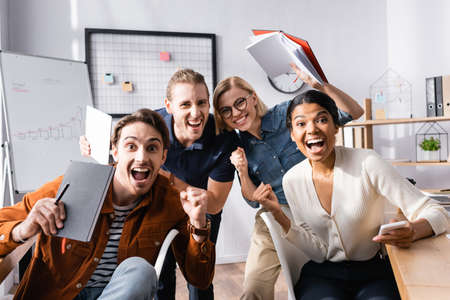excited multiethnic businesspeople shouting while looking at camera and holding notebooks
