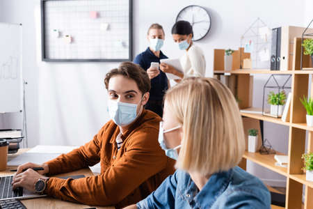 young manager in medical mask looking at colleague while businesspeople talking on blurred backgroundの写真素材