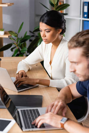 Focused african american woman looking at laptop while sitting at workplace with blurred man on...