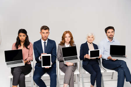 Multiethnic businesspeople showing devices with blank screen while sitting on chairs on gray backgroundの写真素材