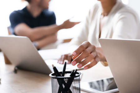Cropped view of hand of african american woman taking pen from holder on desk with blurred man on backgroundの写真素材