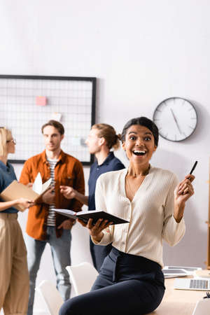 excited african american businesswoman holding notebook and pen near multicultural colleagues...