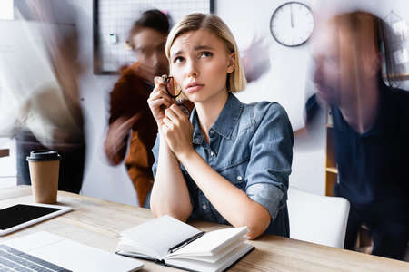 sad businesswoman holding eyeglasses while sitting near notebook in open space office, motion blur