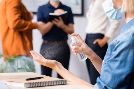 cropped view of businesswoman in medical mask spraying antiseptic on hands near colleagues on...