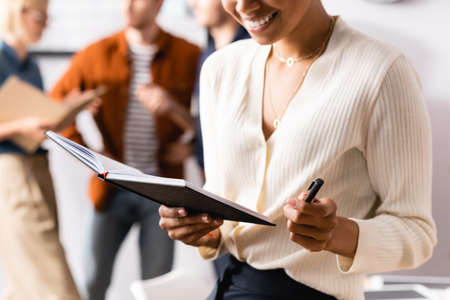 cropped view of african american businesswoman holding notebook and pen near colleagues on blurred backgroundの写真素材