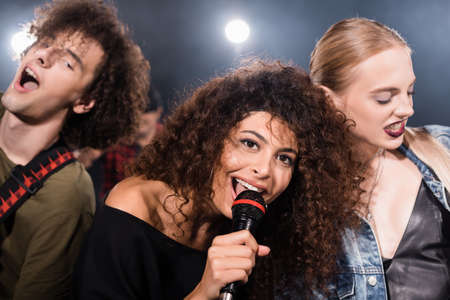 Happy rock band vocalist with microphone singing near musicians with backlit on blurred backgroundの写真素材