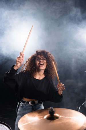 Female drummer with closed eyes shouting, while playing on drum plate with smoke on black backgroundの写真素材