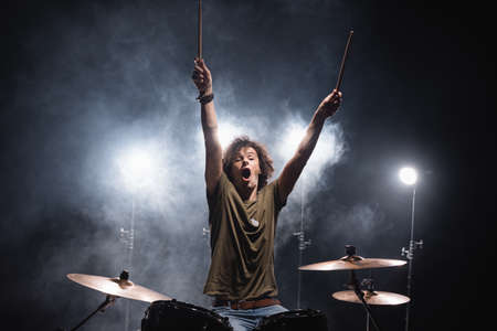 Shouting musician with hands in air sitting at drum kit with smoke and backlit on backgroundの写真素材