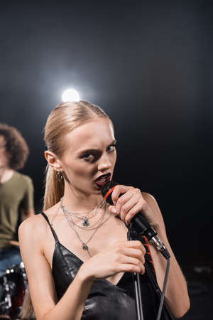 Angry blonde woman singing while touching microphone rack with backlit and blurred drummer on...