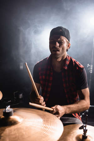 Rock band musician holding drumsticks and playing on drums with smoke on black
