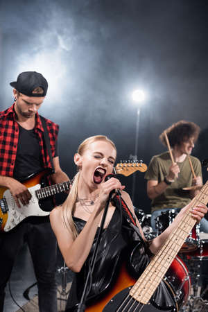 KYIV, UKRAINE - AUGUST 25, 2020: Female vocalist of rock band shouting in microphone near guitarist with backlit and blurred drummer on backgroundのeditorial素材