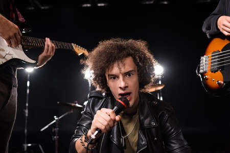 KYIV, UKRAINE - AUGUST 25, 2020: Curly vocalist with microphone looking at camera near musicians...