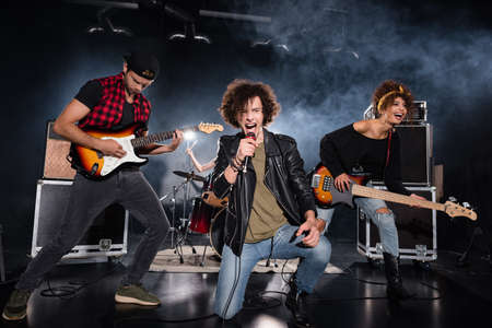 KYIV, UKRAINE - AUGUST 25, 2020: Curly vocalist shouting in microphone while sitting on knee near...