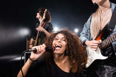KYIV, UKRAINE - AUGUST 25, 2020: Happy curly woman with microphone singing near rock band guitarists with back light on blackのeditorial素材