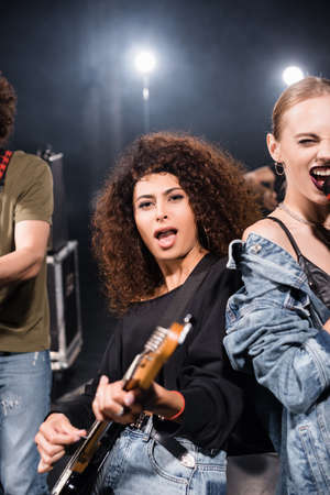 KYIV, UKRAINE - AUGUST 25, 2020: Curly female musician with open mouth playing electric guitar standing near vocalist with backlit on blurred backgroundのeditorial素材
