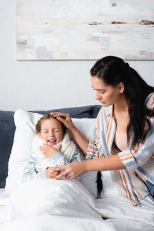 mother touching head of sick daughter coughing in bed with closed eyesの写真素材