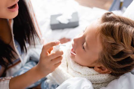 sick girl with open mouth and closed eyes near mother applying throat sprayの写真素材