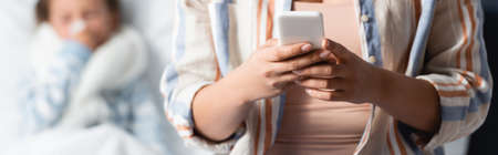 mother messaging on smartphone near sick daughter lying in bed on blurred background, bannerの写真素材