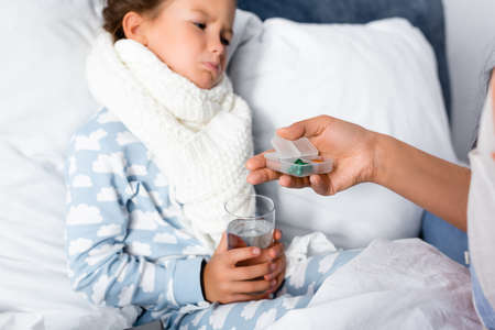 mother giving pills to sad, diseased daughter holding glass of water on blurred backgroundの写真素材