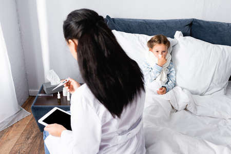 doctor looking at thermometer near diseased girl lying in bedの写真素材