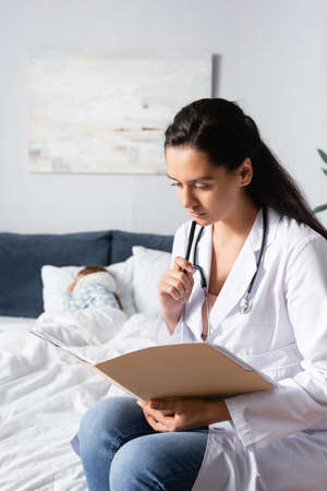 serious doctor looking at outpatient card while diseased child sleeping in bed on blurred backgroundの写真素材