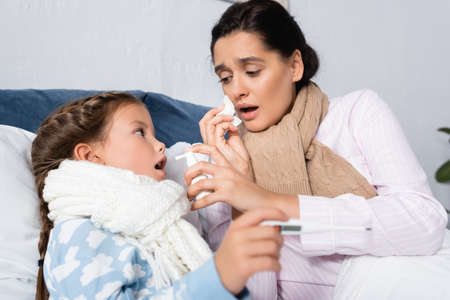 ill woman with runny nose holding throat spray near diseased girl with thermometer on blurred foregroundの写真素材
