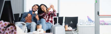 happy bearded man pointing with finger at passport while taking selfie with african american woman in airport, bannerの写真素材