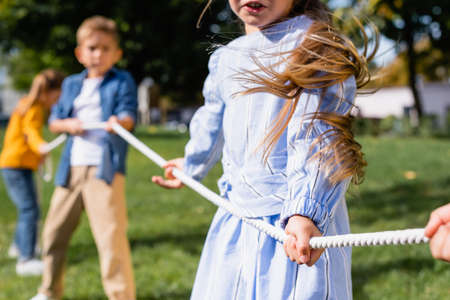Girl holding rope while playing tug of war with friends on blurred background in parkの写真素材