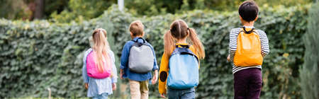 Back view of pupils with backpacks walking near friends on blurred background outdoors, banner