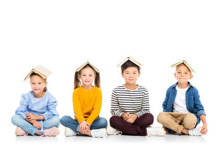 Smiling multiethnic kids with books on heads sitting on white backgroundの写真素材