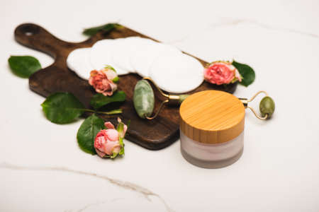 container with cosmetic cream near cotton pads, tea roses, and jade roller on white marble backgroundの写真素材